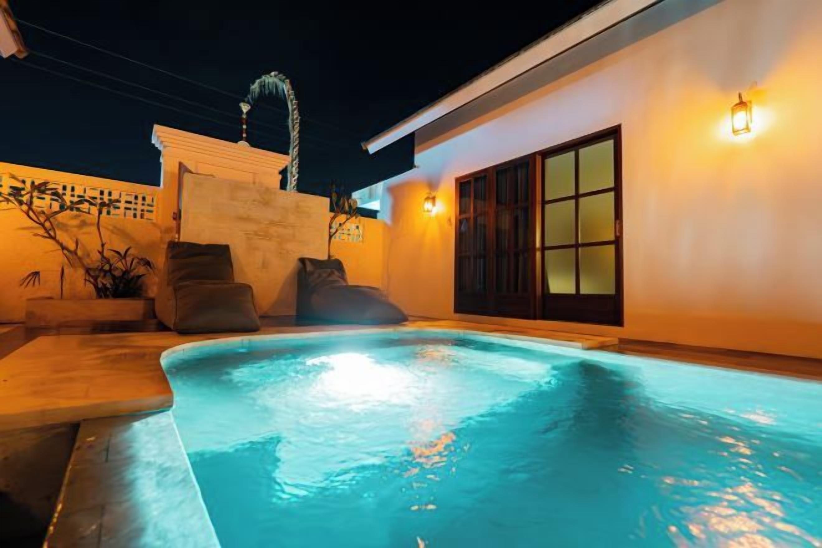 A swimming pool with a tiled deck, a small seating area with cushions, and a building with a sliding glass door.