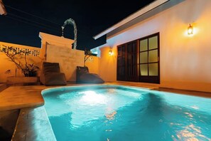 Piscina com deck ladrilhado, um espaço de estar pequeno com almofadas, e um edifício com porta de correr de vidro.