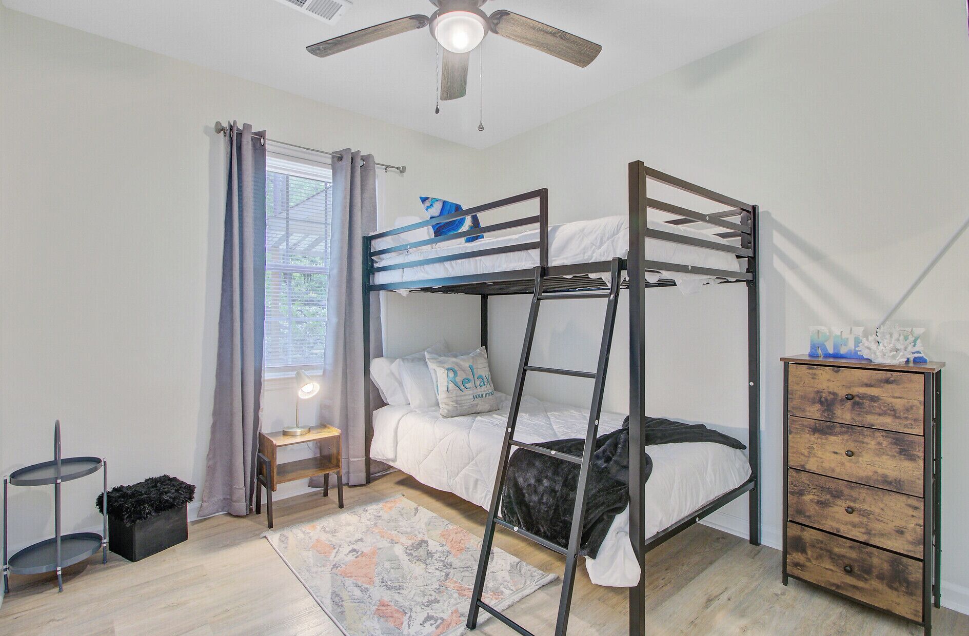 Habitación con literas, ventilador de techo, ventana con cortinas, mesita de noche con lámpara, cómoda y alfombra.