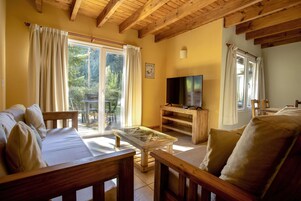 Una sala de estar acogedora con un sofá, una mesa de centro de vidrio, muebles de madera, una televisión y ventanas grandes con cortinas.