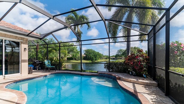 A swimming pool with a clear view of a lake and palm trees, enclosed by a glass structure. - Lakefront Retreat in Prestigious Master Reserve at Lely Resort (Naples)