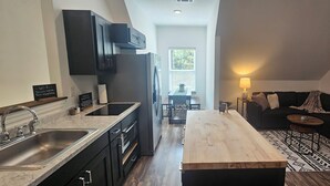 Een moderne keuken met donkere kasten, een houten aanrechtblad en een inbouwoven. Er is een zithoek met een tafel en stoelen, en een woonkamer met een zwarte bank en een kamerbreed tapijt.