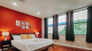 A bedroom with a wooden floor, a bed with white linens, two bedside tables with lamps, a window with curtains, and artwork on the wall. - Prime / 2 King Beds / 65" TV / Jefferson Hospital (Philadelphia)