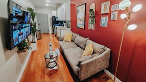 A modern living room with a red accent wall, a grey sofa, and a flat-screen TV displaying NBA highlights. - Prime / 2 King Beds / 65" TV / Jefferson Hospital (Philadelphia)