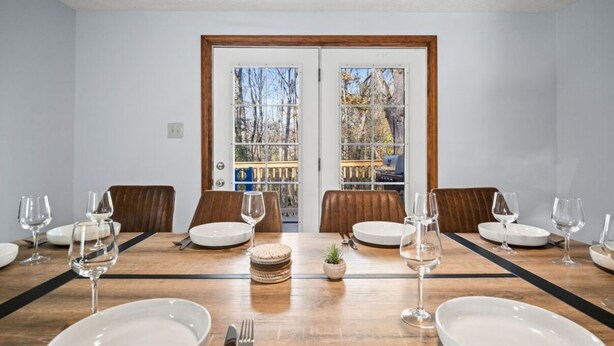 Ein Esszimmer mit einem Holztisch, gedeckt für sechs Personen, zwei Holzstühle und Blick auf eine Terrasse durch eine Glastür.
