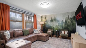 Ein Wohnzimmer mit Ecksofa, einem kleinen Tisch, einer Lampe und einem an der Wand montierten Fernseher.