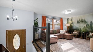Ein Wohnzimmer mit Ecksofa, einer Deckenleuchte, einer Treppe und einem Wandbild von einem Wald.