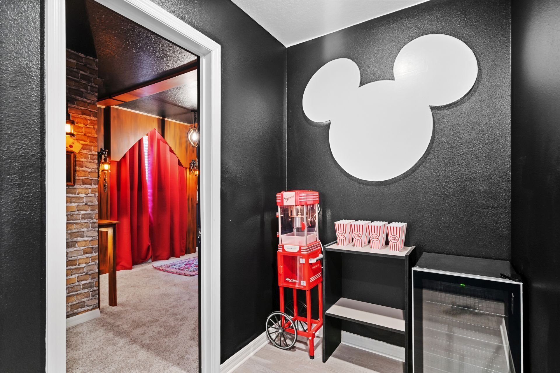 A room with a black wall featuring a large white Mickey Mouse logo, a red popcorn machine, and a popcorn dispenser.