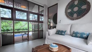 A modern living room with a white sofa, a round wooden coffee table, and a large window with a view of greenery. - Villa Blue Paradise, Villa 22 (Koh Samui)