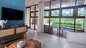 A modern living room with a TV, wooden furniture, a dining table with a cake, and large windows with a view of greenery. - Villa Blue Paradise, Villa 22 (Koh Samui)