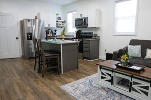 A modern kitchen with a central island, a dining area with a wooden chair, and a living room with a sofa and decorative pillows.