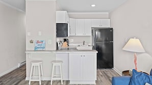 A compact kitchen with white cabinetry, a black refrigerator, and a marble countertop. There are two white stools and a small island. - Steps to Sand! 3rd-Floor North Topsail Beach Condo (North Topsail Beach)
