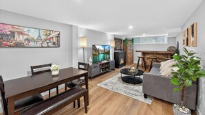 A modern living room with a dining area, a fireplace, and a large painting of a street scene. - Bright & Modern 2BR Basement Suite w/ Rustic Bar & Private Entrance (Kingston)