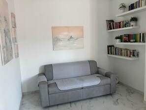 Ein graues Sofa mit einer gemusterten Decke, ein Wandbild und ein Bücherregal mit Büchern und Pflanzen.