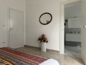 A bedroom with a bed, a white door, a wall-mounted mirror, and a bathroom visible through an open door.