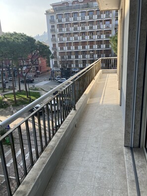 A balcony with a metal railing overlooking a street with cars and a multi-story building in the background.