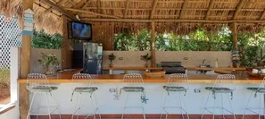 Eine Strandbar mit einem Reetdach, weißen Metallstühlen und einem an der Wand befestigten Fernseher.