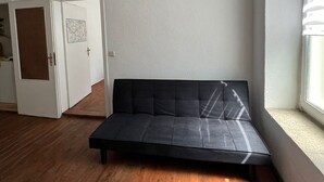 Une pièce avec un canapé noir, un plancher en bois, une porte blanche et une fenêtre avec des stores.
