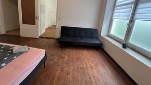 Une chambre à coucher avec un lit, un canapé noir, un plancher en bois, une fenêtre avec des stores et une porte donnant sur une autre pièce.