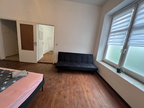 A bedroom with a bed, a black sofa, a wooden floor, a window with blinds, and a door leading to another room.