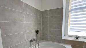 Une salle de bain moderne avec une baignoire sur pieds, des murs recouverts de carreaux gris et une fenêtre avec des stores.