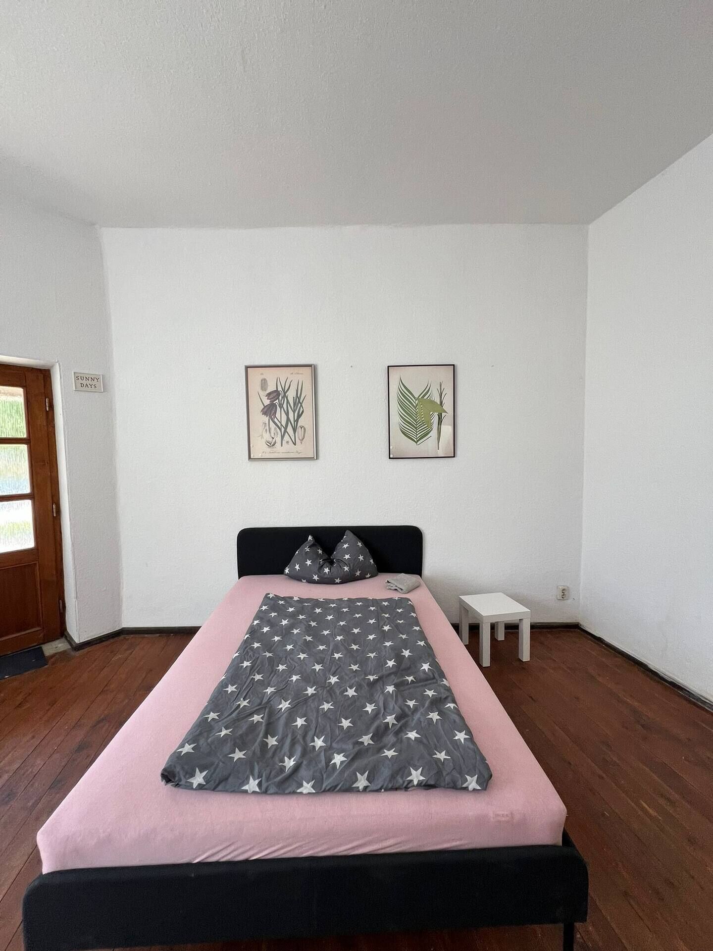 A bedroom with a bed, a small bedside table, and two framed botanical prints on the wall.