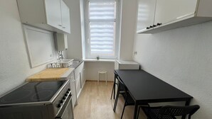 Une cuisine de taille réduite, avec des meubles blancs, une cuisinière, un évier et une fenêtre équipée de stores.