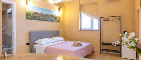 A bedroom with a bed, a window, a painting, a towel, a bench, and a flower vase.