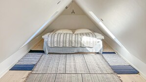 Quarto loft com teto inclinado, cama com manta estampada e dois tapetes no chão.