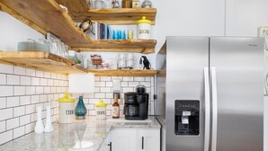 Una cocina con un refrigerador de acero inoxidable, una cafetera y un dispensador de agua. Hay repisas de madera sobre la encimera con diversos utensilios de cocina.