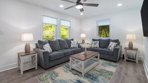 Ein modernes Wohnzimmer mit zwei grauen Sofas, einem Holztisch und zwei Nachttischen mit Lampen. Es gibt einen Deckenventilator und große Fenster mit Jalousien.