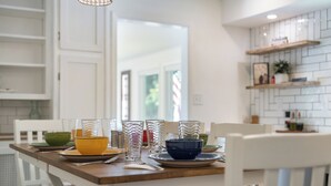 Ein Esstisch mit unterschiedlichem Glas und Schüsseln, weiße Stühle und eine Küche mit weißen Schränken und einem Regal.