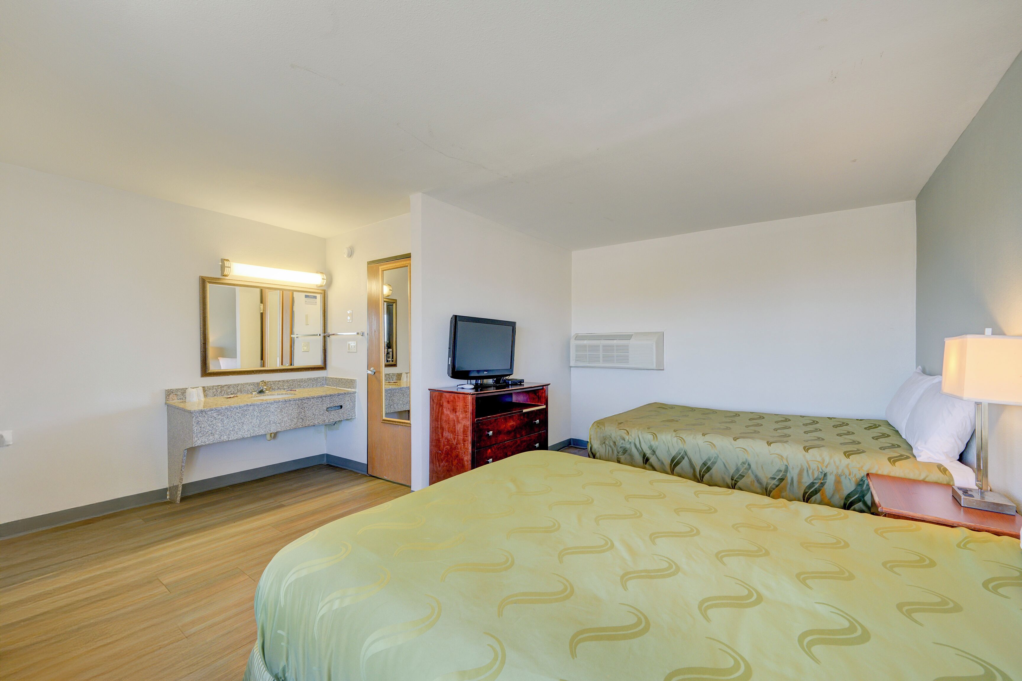 Habitación de hotel con dos camas, televisor, cómoda, espejo y baño con lavamanos y espejo.