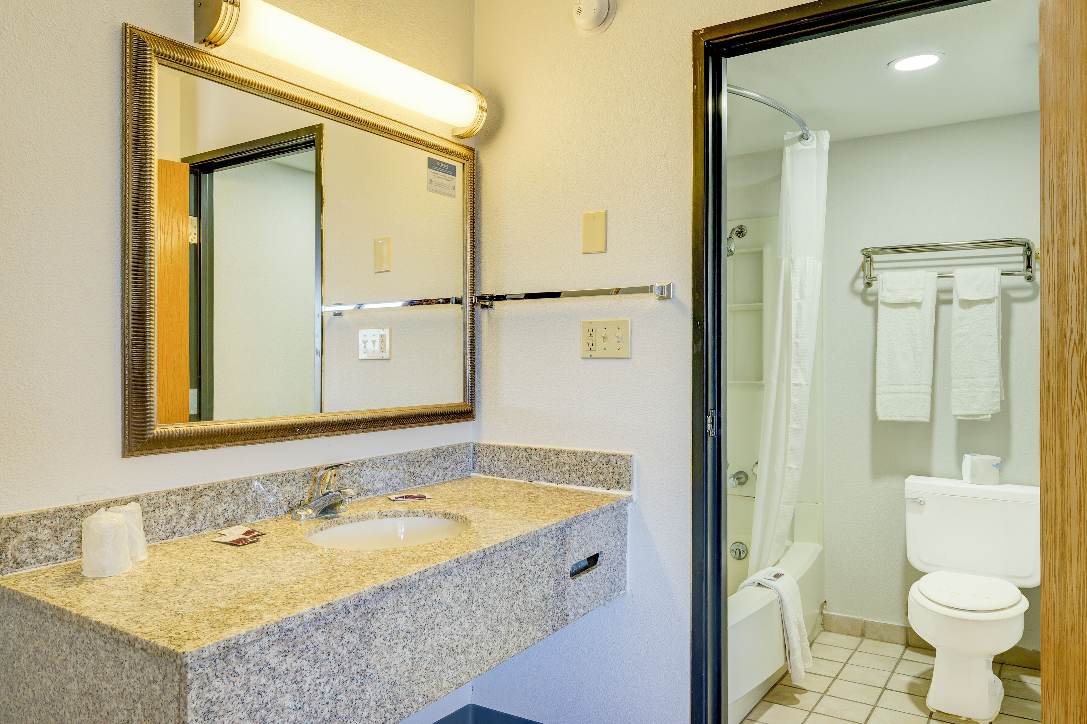 Een badkamer met een wastafel met granieten blad, een spiegel met gouden lijst, een handdoekrek met witte handdoeken en een wit toilet.