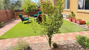 A backyard with artificial grass, a wooden table, two green chairs, a small tree, and a brick wall.