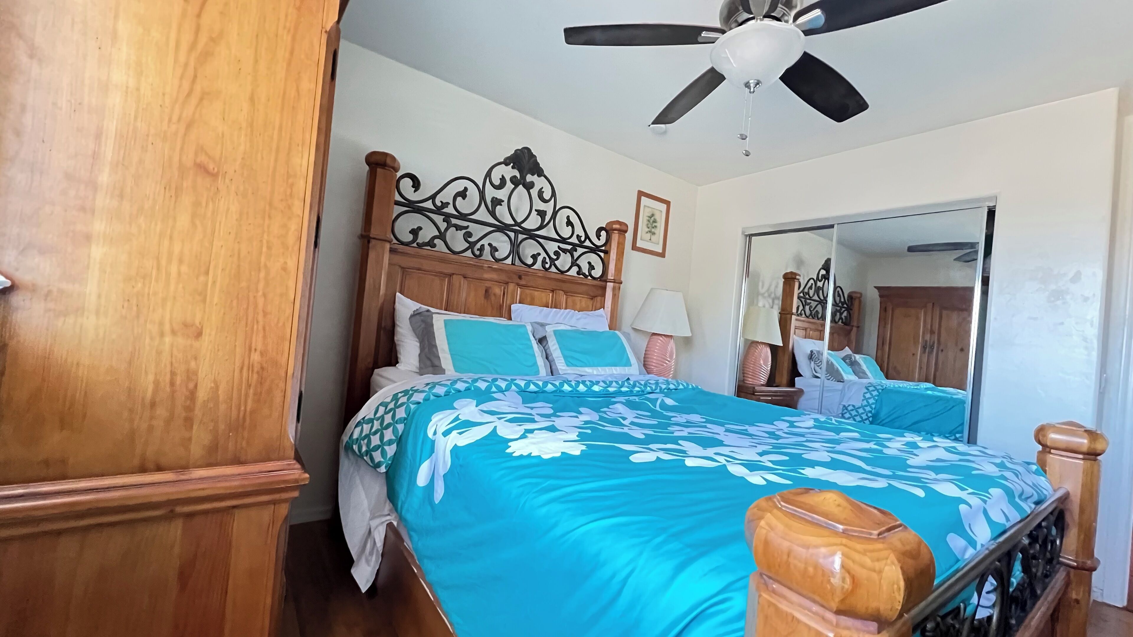 A bedroom with a wooden headboard, a bed with a blue floral bedspread, a ceiling fan, and a mirror.