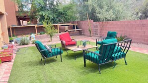 Ein Garten mit Terrassenbereich, grünem Kunstrasen und Gartenmöbeln, darunter zwei Stühle und ein Tisch.