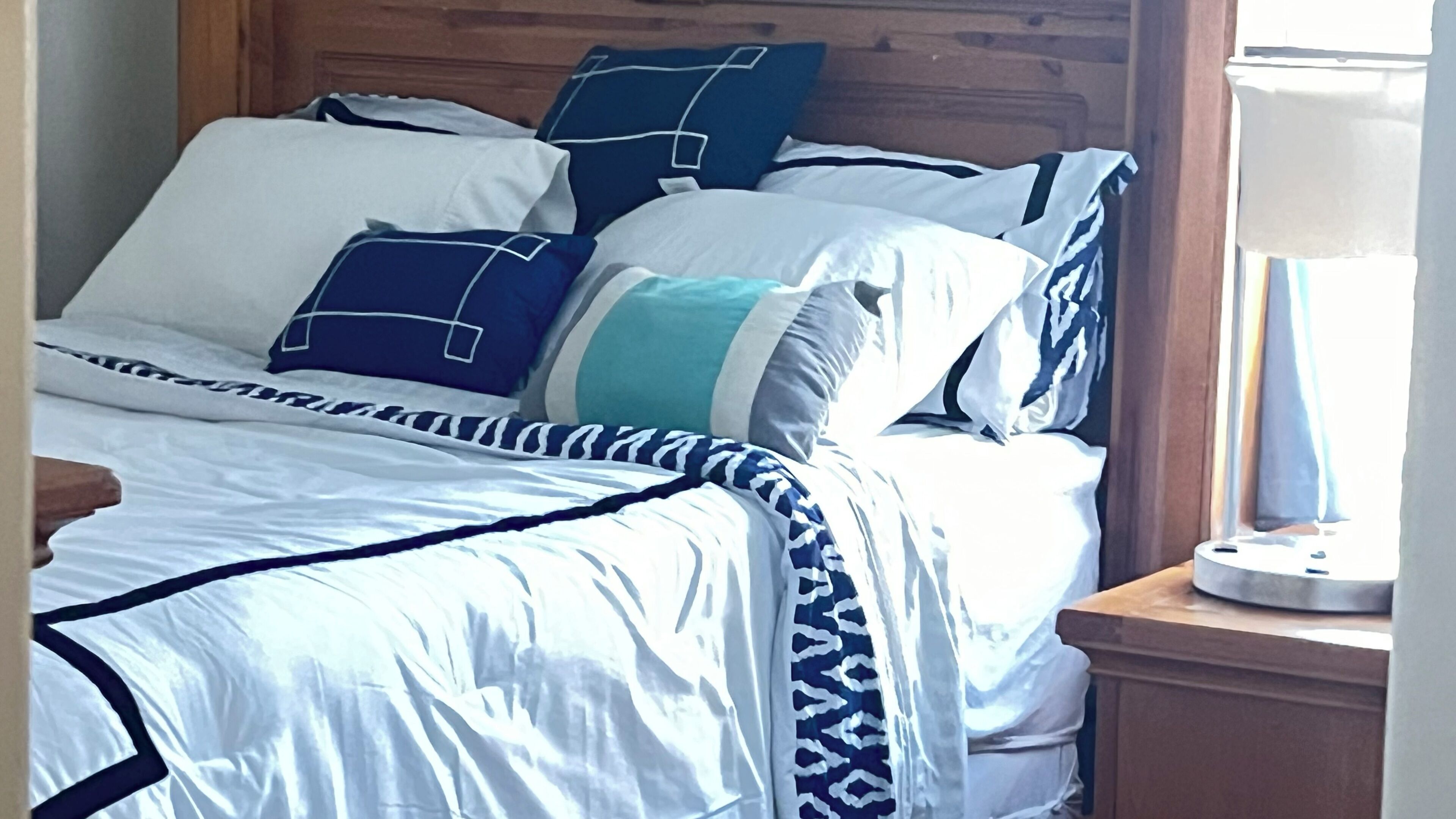 A bed with a wooden headboard, white and blue bedding, and a bedside table with a lamp.
