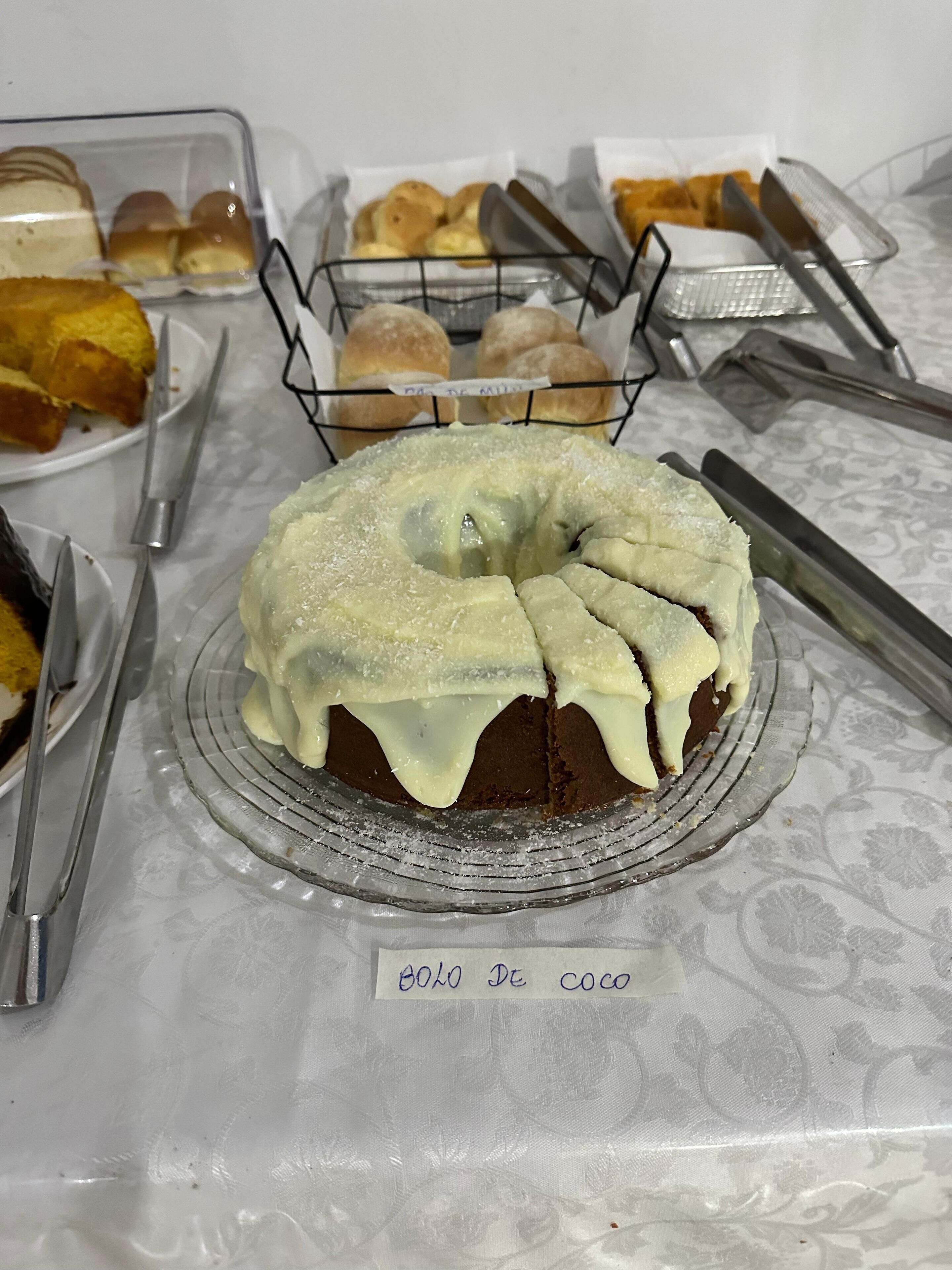 Un pastel con una etiqueta que dice 'BOLO DE COCO' sobre una mesa con diversos productos horneados y utensilios.