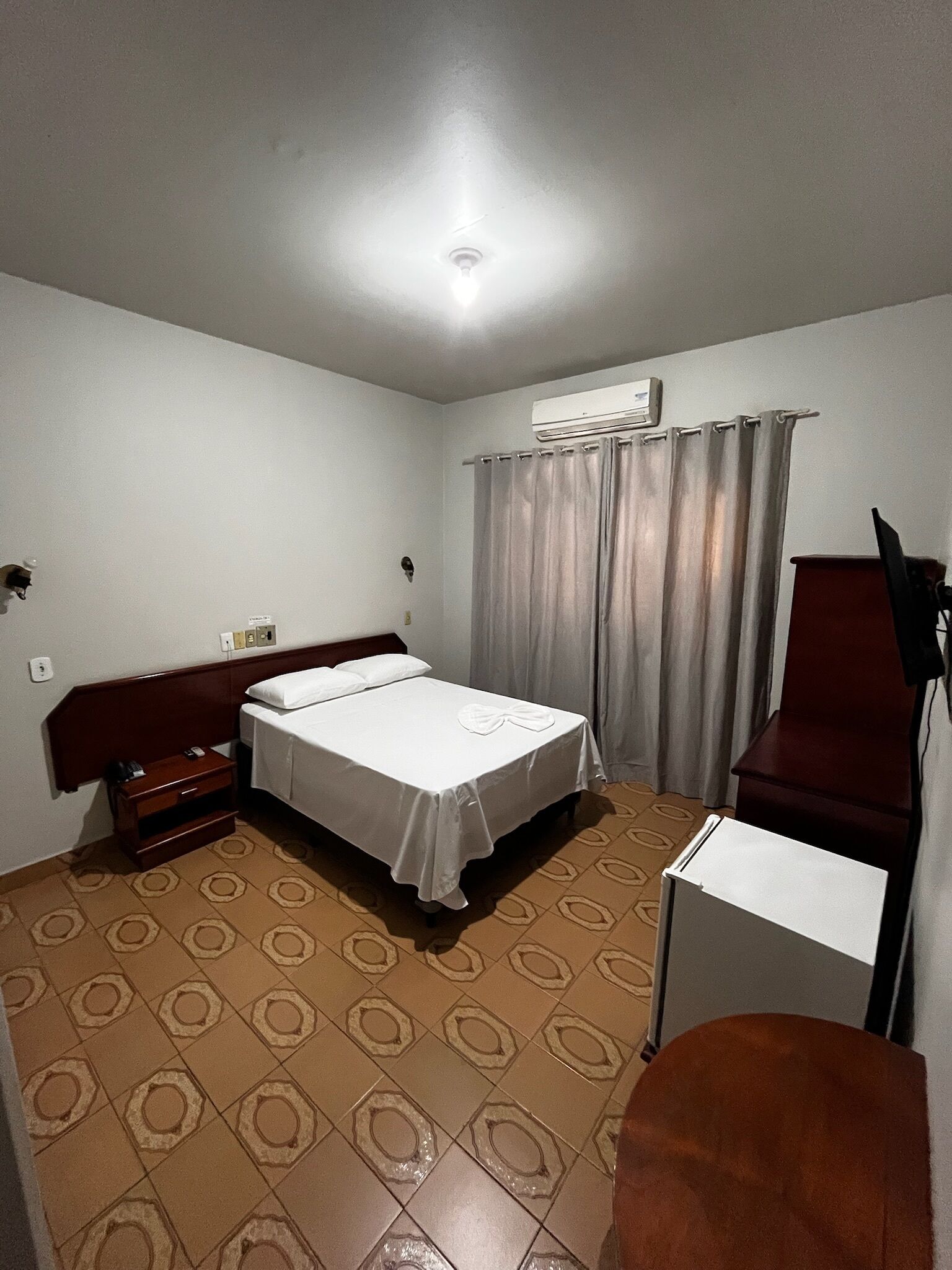 A small, well-lit room with a single bed, a nightstand, a desk with a chair, and a wall-mounted air conditioner.