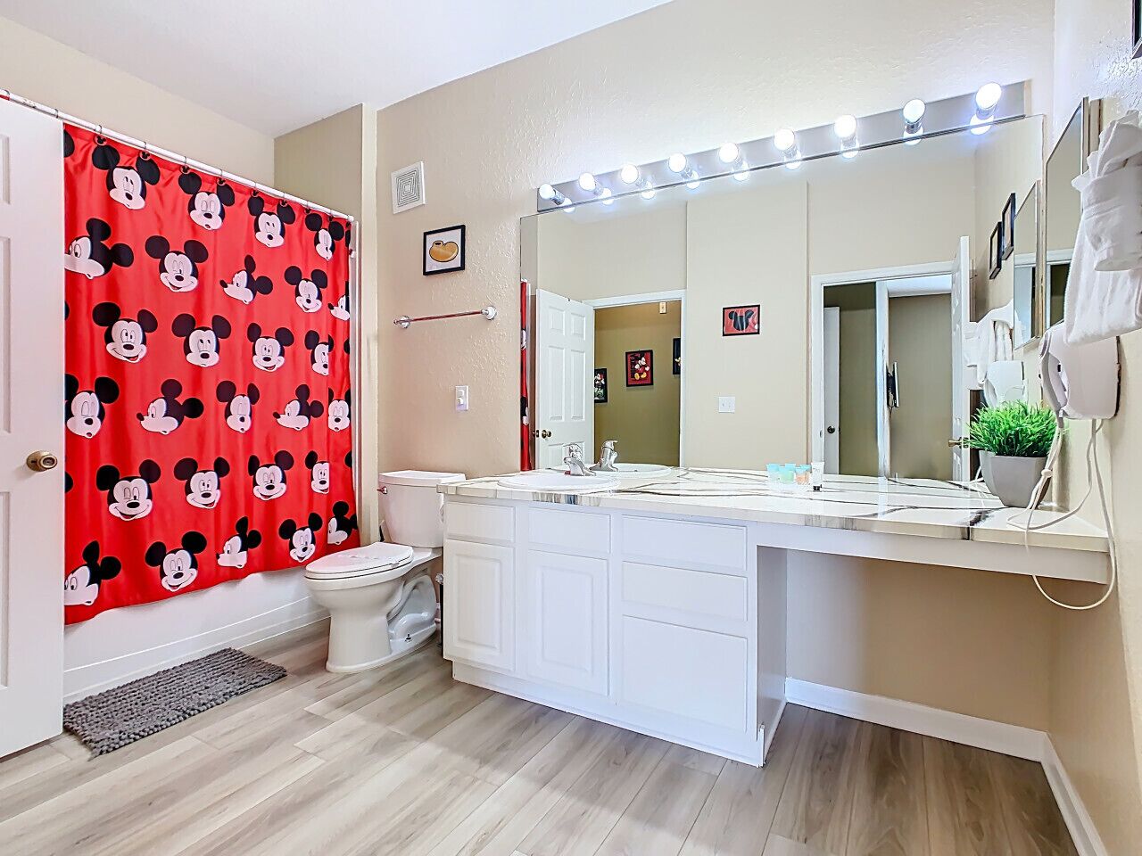 Une salle de bain avec un rideau de douche à l’effigie de Mickey Mouse, des toilettes blanches, un meuble-lavabo doté d’un grand miroir et un évier.