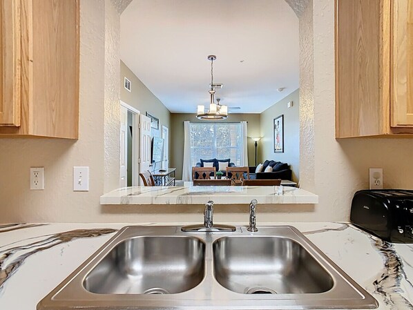 A kitchen with two stainless steel sinks, a marble countertop, wooden cabinets, and a view into a living area with a sofa and a dining table.