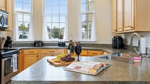 A kitchen with wooden cabinets, a granite countertop, a wine bottle, a wine glass, a cutting board with fruits, and a cookbook.