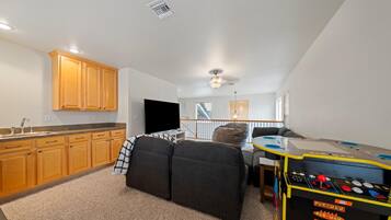 A compact living space with a kitchenette, a flat-screen TV, a bean bag chair, a gaming table, and a staircase leading to an upper level.
