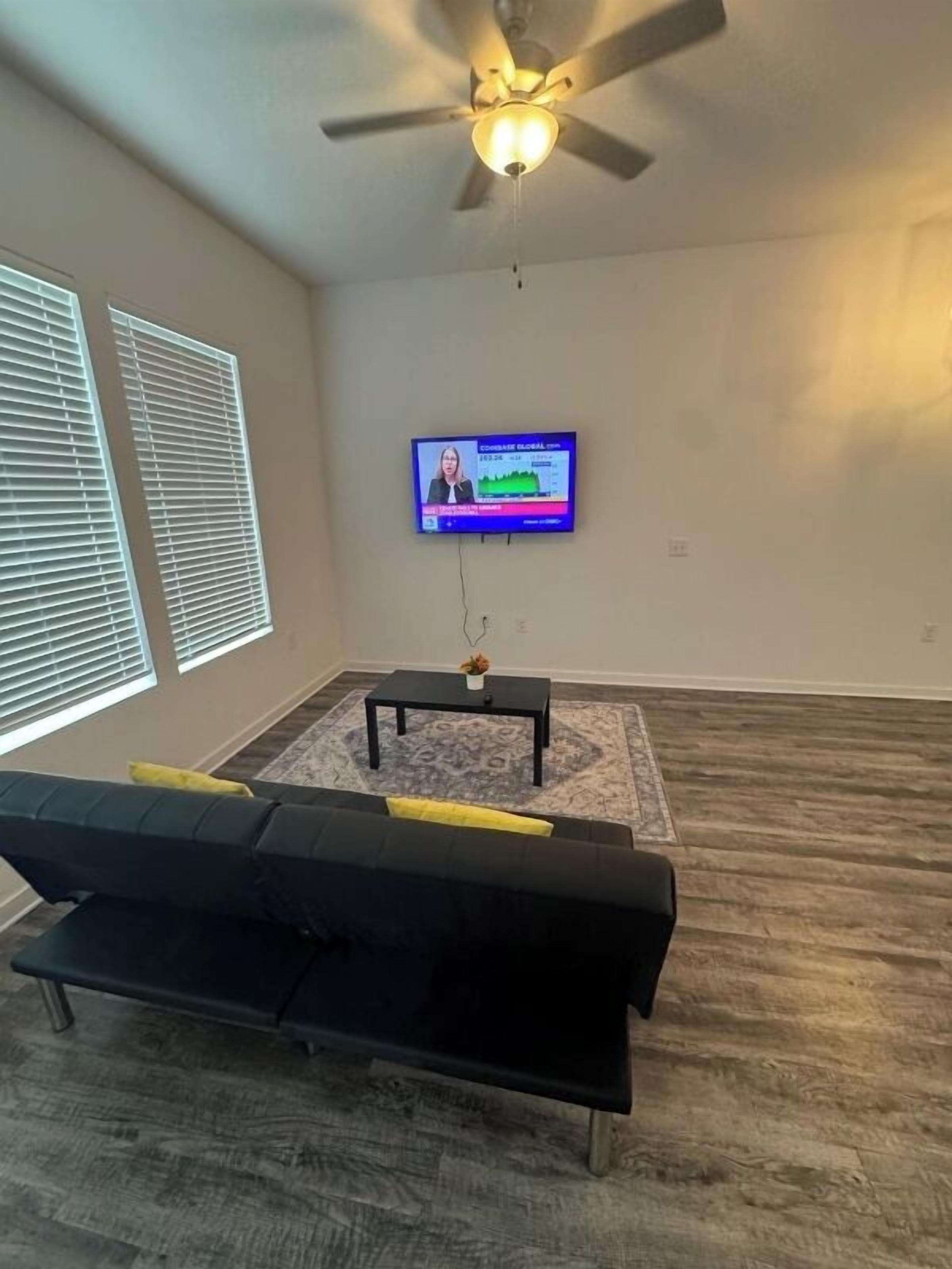 A living room with a black sofa, a small table with a vase, a ceiling fan, and a TV mounted on the wall.