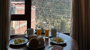 A breakfast table with plates of food, glasses of orange juice, and a mug, set against a window with a view of a mountainous landscape. - JUAN PLACIDO Hospedaje (Cusco)