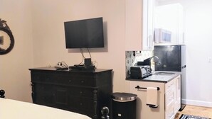 Ein kleines Zimmer mit einem Bett, einem Schrank, einem Fernseher, einer kleinen Küche und einem Mülleimer.