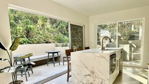 A modern kitchen with a marble island, large windows, and a view of greenery. - The Modern Kailua Beach House (Kailua)