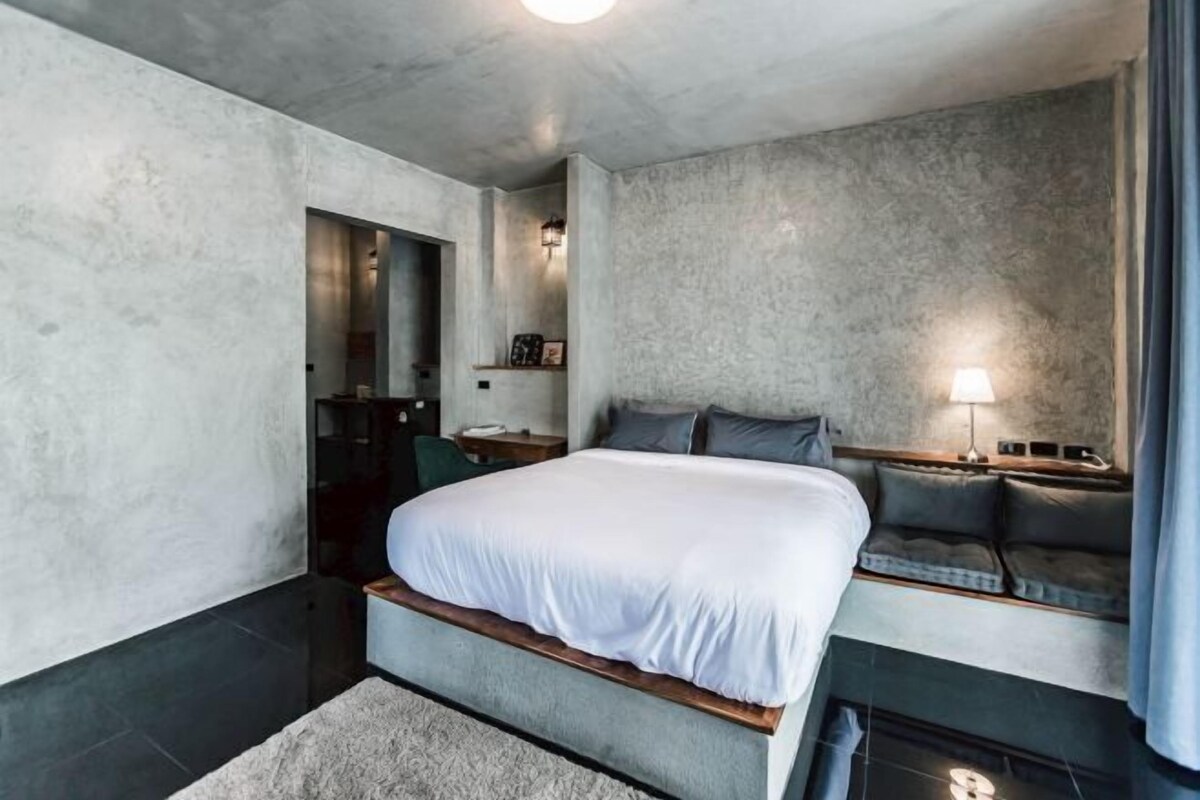 Een moderne slaapkamer met een groot bed, een zithoek met kussens, een klein tafeltje en een wandlamp.