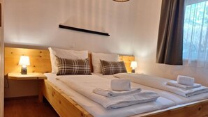 A double bed with white linens and two pillows, a wooden headboard, bedside lamps, and a window with curtains.